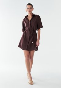 Brun kortærmet kjole med bælte ved taljen, med krave og en let A-formet silhuet, lavet af et tekstureret stof.