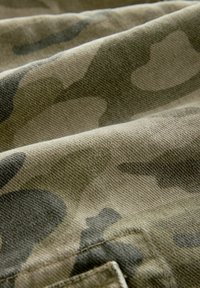 Camo-mønstret stof i nuancer af grøn og brun, med en tekstureret overflade og synlige syninger. Blødt, alsidigt materiale.