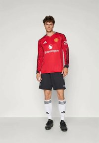 Maglia da calcio a maniche lunghe rossa con strisce nere e logo, abbinata a pantaloni corti neri. Calze ginocchiali bianche con strisce nere e scarpe da calcio nere.