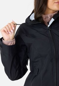 Sort vandtæt jakke med lynlåslukning foran, justerbar hætte og tekstureret stof. Synlige håndlommer med lynlåse, indre manchetter vises.
