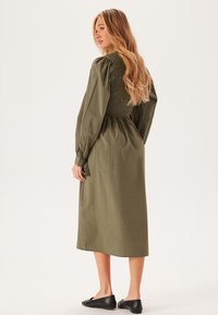 Robe longue à manches longues de couleur vert olive avec un corsage à smocks, une jupe fluide et une texture subtile ; portée avec des chaussures plates noires.
