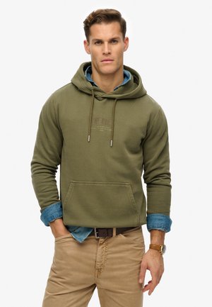 Felpa verde oliva con tasca a marsupio e cordini. Indossa una camicia in denim blu chiaro sotto e pantaloni chinos beige.