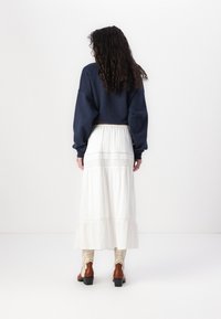 Kvinna med lockigt brunt hår som bär en marinblå tröja, vit midikjol med spetsdetaljer och beige och bruna cowboyboots, stående med ryggen vänd.