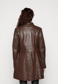 Brun läderjacka med en figursydd silhuett och spetsig krage. Har sömdetaljer och sidoremmar. Slät textur och medellång design.