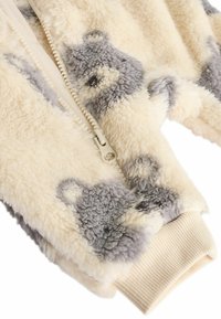 Jakna krem boje sa zipom, mekane i plišaste teksture, sivo-smedastih uzoraka medvjedića i rebrastim manžetama. Zipper je bež boje i glatka.
