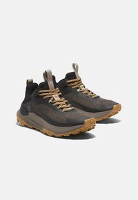 Zapatillas de trail con una parte superior de malla negra, acentos de goma, cordones en tonos tierra y una suela texturizada en marrón para mejorar la tracción.