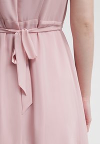 Gros plan de l'arrière d'une robe rose clair avec une ceinture en tissu nouée à la taille et un bras visible sur le côté droit.