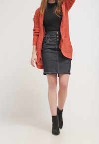 Falda lápiz de mezclilla oscura con cintura alta y tres botones decorativos, combinada con una camiseta negra ajustada y un cárdigan coral de punto abierto. Botines negros.