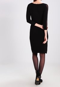 Mustat bodycon-mekko, jossa on teksturoitu kangas, kolme neljännesosaa hihahuulia läpikuultavilla yksityiskohdilla ja polvipituinen malli. Esitetty mustien tiheiden sukkahousujen kanssa.