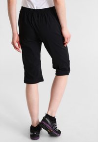 En person som bär svarta knälånga shorts och svart-lila Merrell träningsskor, stående mot en enkel ljus bakgrund.