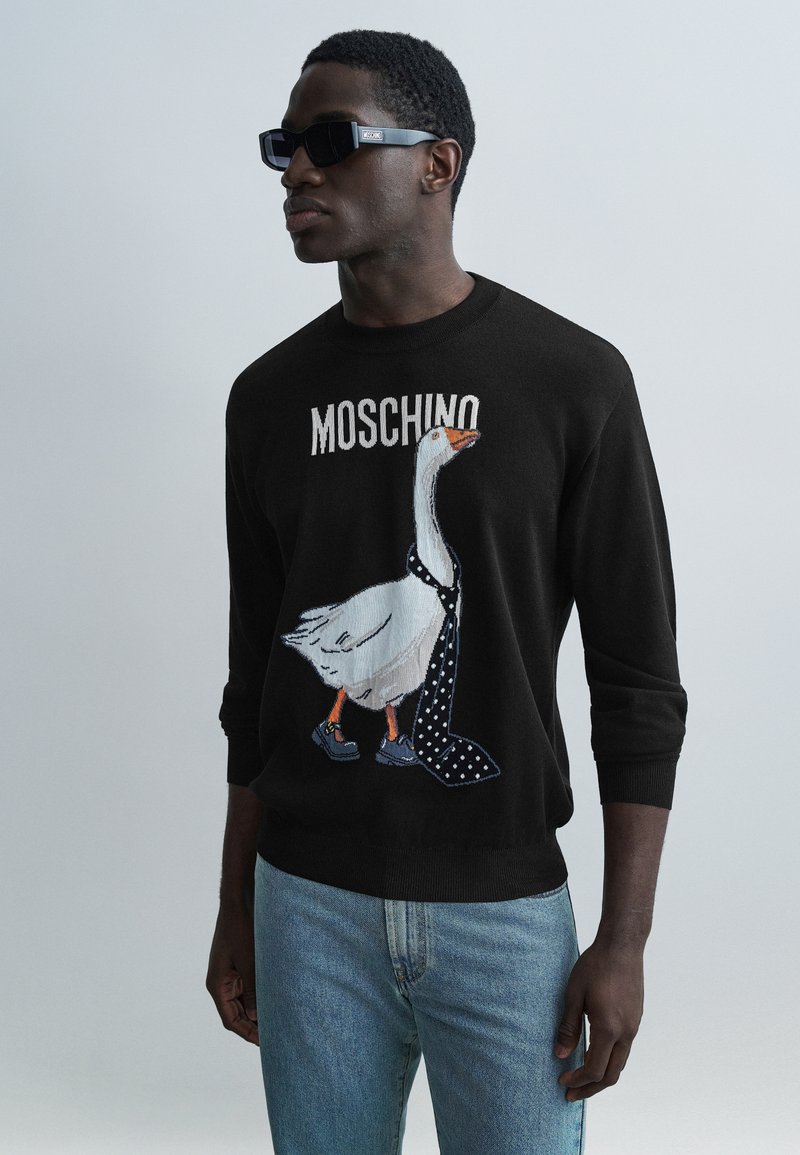 Schwarzer Pullover mit weißem Gänsemotiv, das eine gepunktete Krawatte und Schuhe trägt. Über der Grafik befindet sich der Schriftzug „Moschino“.