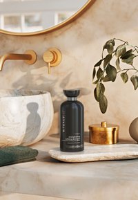 Bottiglia nera dello "Shampoo Rinforzante al Caffeina Rituals Homme" su un vassoio di marmo chiaro, con un bacino di marmo e un contenitore con accenti dorati nelle vicinanze.