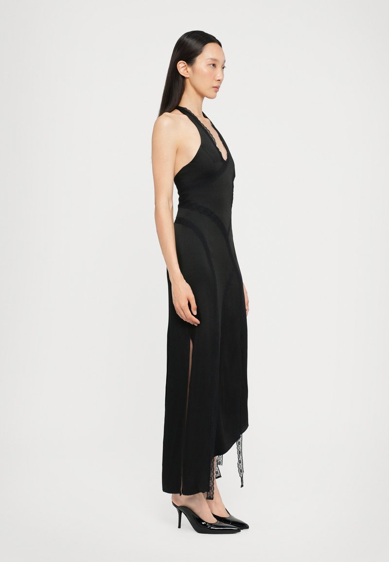 Black halter neck dress with a fitted silhouette, side slits, lace trim detailing, and a sheer underlay at the hem, paired with black heels.