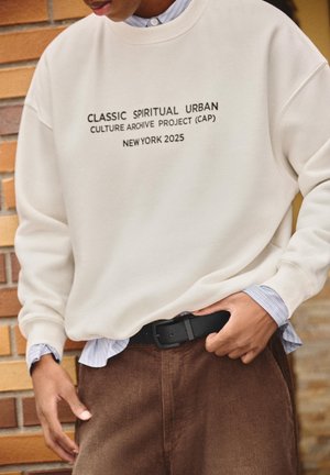 Persoană purtând un hanorac alb cu textul „Classic Spiritual Urban Culture Archive Project (CAP) New York 2025”, pantaloni maro și o curea neagră.