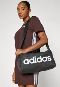 Black fabric bag with a large white "adidas" logo, rectangular shape, dual handles, and an adjustable shoulder strap.