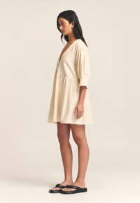 Korte beige jurk met een diepe V-hals, puffmouwen en een gathered taille. Draagt een paar zwarte slippers. Soepele, lichtgewicht stof.