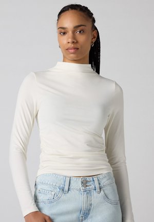 Maglietta a maniche lunghe e collo alto in tessuto color crema con una superficie morbida e dettaglio arricciato in vita, abbinata a jeans in denim azzurro chiaro.