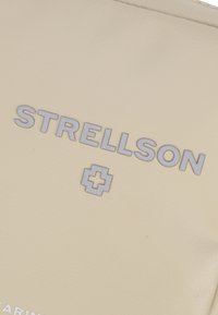 Il materiale color crema presenta il testo in rilievo "STRELLSON" e un logo a forma di croce argentata, con una superficie liscia e texture.