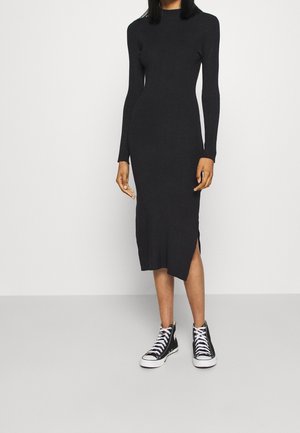 Schwarzes, langärmliges Rippkleid mit hohem Ausschnitt und seitlichem Schlitz, kombiniert mit schwarzen High-Top Sneakers. Der Stoff hat eine glatte Textur.
