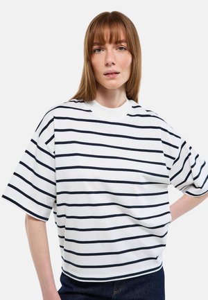 Jeune femme aux cheveux châtain clair et raides portant une chemise blanche oversize à rayures horizontales noires, debout devant un fond uni.