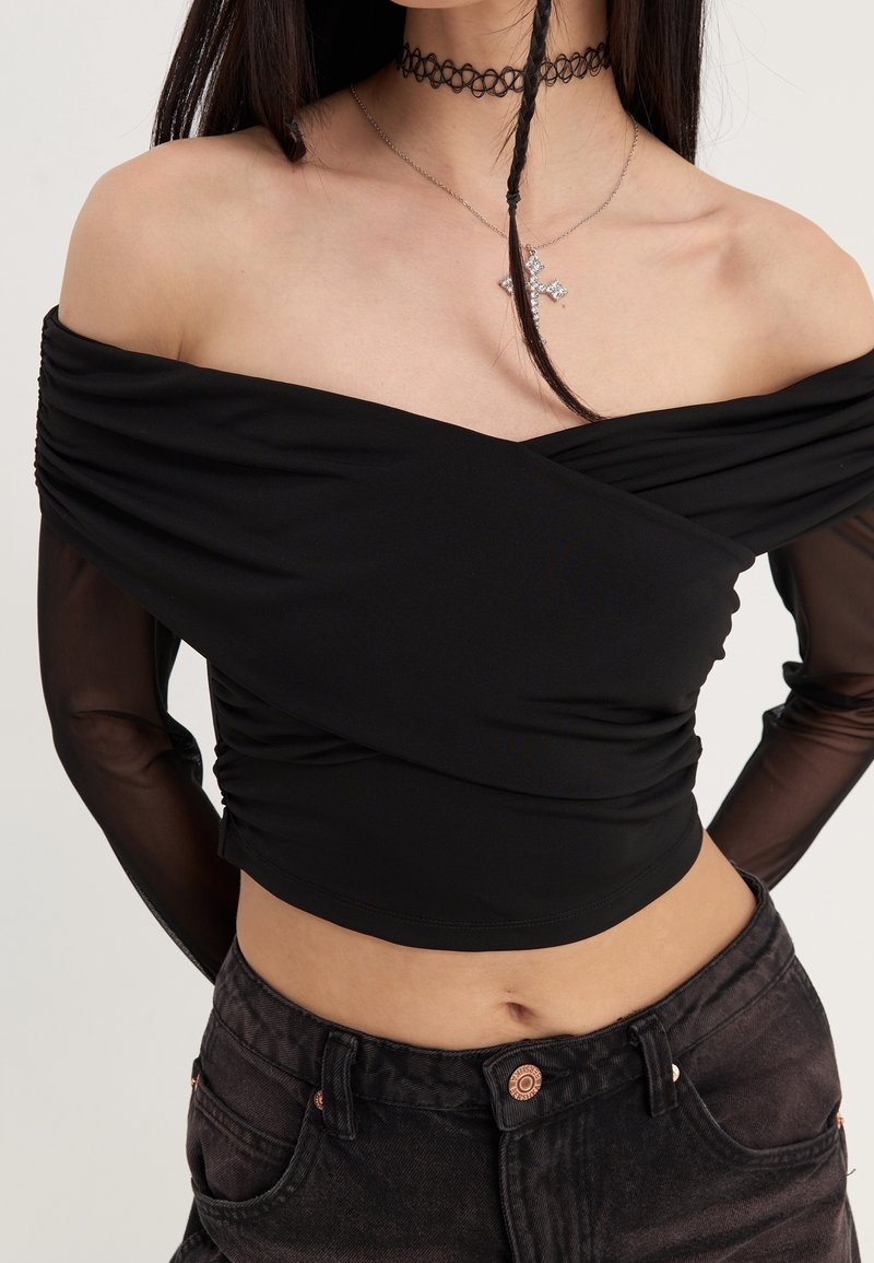 Femme portant un crop top noir épaules dénudées avec manches longues transparentes, un jean noir taille haute, un collier ras de cou noir et un pendentif croix argenté.