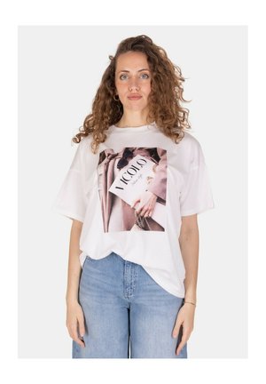 Giovane donna con capelli ricci che indossa una maglietta bianca con stampa fotografica e jeans azzurri chiari, in piedi davanti a uno sfondo semplice.