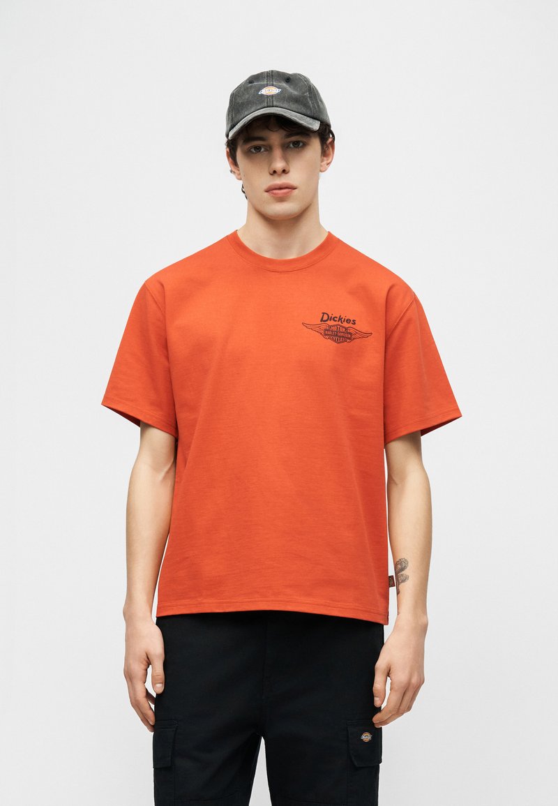 Junger Mann mit verwaschener schwarzer Mütze, orangefarbenem Dickies-T-Shirt mit Logo auf der Brust und schwarzen Cargo-Hosen mit Seitentaschen.