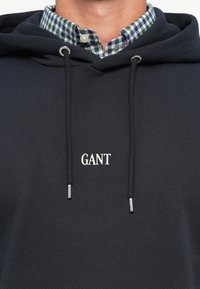 Hombre vistiendo una camisa de cuadros con botones debajo de una sudadera azul marino con el logo "GANT" y los cordones visibles alrededor del cuello.
