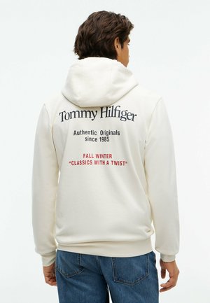 Mężczyzna w białej bluzie z kapturem, z napisem "Tommy Hilfiger" i hasłem na jesień i zimę na plecach, w połączeniu z niebieskimi dżinsami.