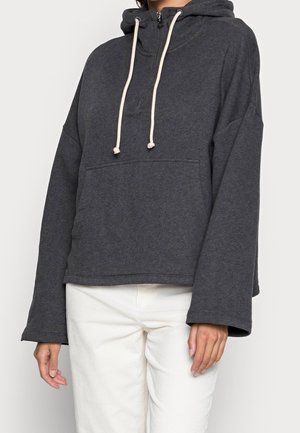 Vrouw draagt een donkergrijze capuchontrui met witte trekkoorden en losse mouwen, gecombineerd met een witte broek, tegen een effen achtergrond.