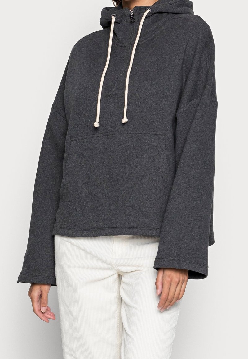 Femme portant un pull à capuche gris foncé avec des cordons blancs et des manches larges, associé à un pantalon blanc sur un fond uni.