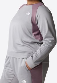 Felpa sportiva grigia con dettagli viola sulle spalle, tessuto testurizzato e logo. Indossata con pantaloni coordinati, con una mano in tasca.