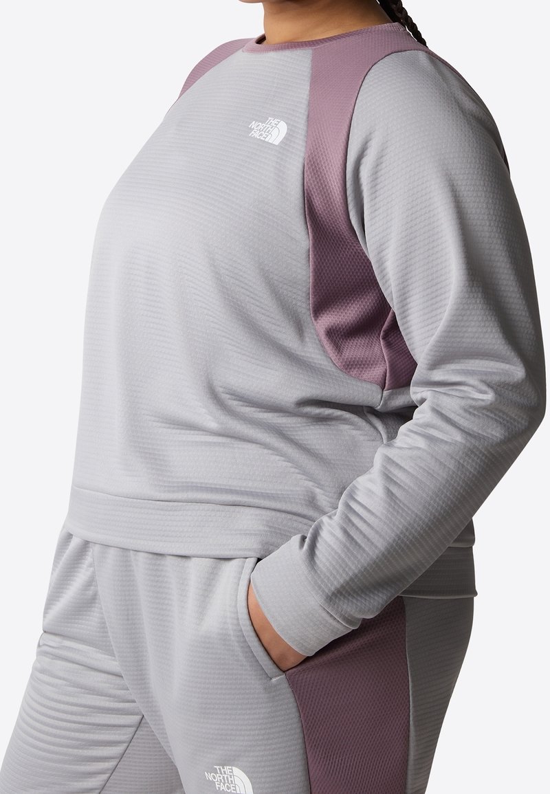 Felpa sportiva grigia con dettagli viola sulle spalle, tessuto testurizzato e logo. Indossata con pantaloni coordinati, con una mano in tasca.