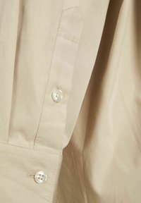 Camicia beige con bottoni, realizzata in cotone liscio, con colletto a punte e un singolo bottone in madreperla sul polsino.