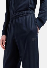 Pantaloni jogger blu navy con vita elastica, tasche laterali e un discreto logo EA7 sulla gamba. Tessuto liscio con una morbida texture.