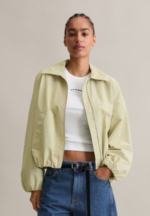 Junge Frau mit geflochtenem Haar trägt eine hellbeige Reißverschlussjacke, ein weißes kurzes Top mit Schriftzug und hochtaillierte blaue Jeans mit schwarzem Gürtel.