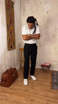 Άνδρας με κότσους, ντυμένος με λευκό μπλουζάκι και μαύρο παντελόνι, στέκεται με σταυρωμένα τα χέρια σε δωμάτιο με ξύλινο πάτωμα, δερμάτινη τσάντα, καρέκλα και υφαντό τοίχου.