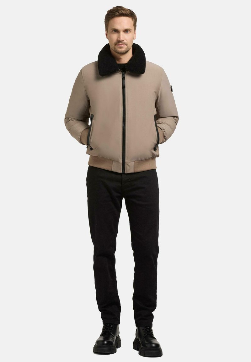 Beige bomberjakke med svart kunstpelskkrage, glidelås og ribbet kant. Brukt med svarte bukser og svarte støvler, stående rett.