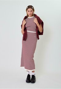 Prugasta pletena haljina u bordo i bijeloj boji, uskog kroja s rebrastom teksturom. Burgundski pulover složen preko ramena; crne cipele.