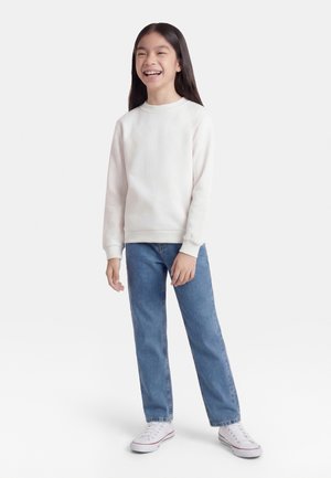 Ung person står og smiler, iført en hvit genser, blå jeans og hvite joggesko på en ensfarget bakgrunn.