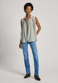 Camicia senza maniche, verde chiaro, con bottoni e colletto, dettagli arricciati sul davanti, abbinata a jeans blu e stivaletti neri alla caviglia.
