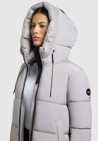 Jachetă puf gri deschis cu o glugă mare, închidere cu fermoar și buzunar frontal. Prezintă secțiuni cusute și un patch cu logo pe mânecă.