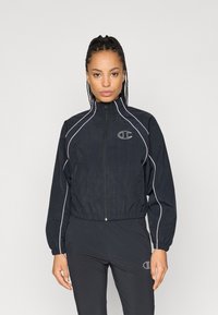 Chaqueta cortavientos negra con un cuello alto, que presenta ribetes blancos y un pequeño logo en el pecho. Ligera, con una cinturilla y puños elásticos.