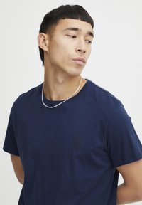 Temno modra bombažna majica s krožnim izrezom in kratkimi rokavi; vsebuje subtilno embosirano grafiko na prsih. Gladka tekstura.