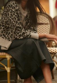 Cardigan à motif léopard gris avec des manches côtelées, porté sur une jupe en cuir noir. La jupe a une texture lisse et tombe en dessous des genoux.