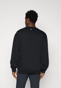 Melnas kokvilnas sporta krekls ar apaļu kaklu, garām piedurknēm un ribotām aprocēm. Aizmugurē, tuvāk kakla līnijai, ir balts logo.