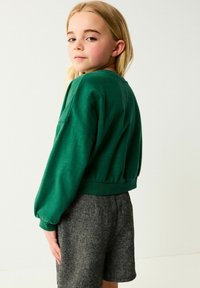 Grüner Baumwollpullover mit weiten Ärmeln und elastischem Saum, kombiniert mit grauen Shorts mit strukturierter Hahnentritt-Optik.