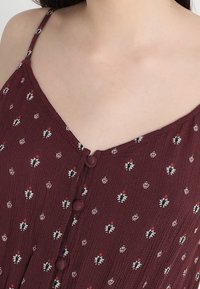 Robe bordeaux à fines bretelles, avec un décolleté en V et de petits boutons sur le devant. Motif floral avec des accents rouges et noirs.