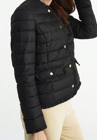 Černá péřová bunda s vypasovaným střihem, zvlněným lemem a zlatými tlakovýma knoflíky. Obsahuje postranní kapsu a horizontální prošívanou texturu.