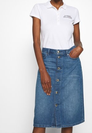 Femme portant un polo blanc à manches courtes avec le logo "Tommy Hilfiger" et une jupe en denim bleu à boutons sur le devant, longueur genou.